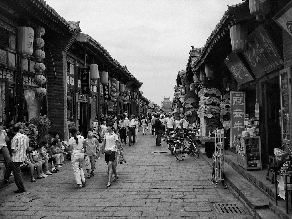 Black and white photograph of a crowded traditional Chinese street lined with historic buildings, lanterns, and shops with pedestrians and bicycles.