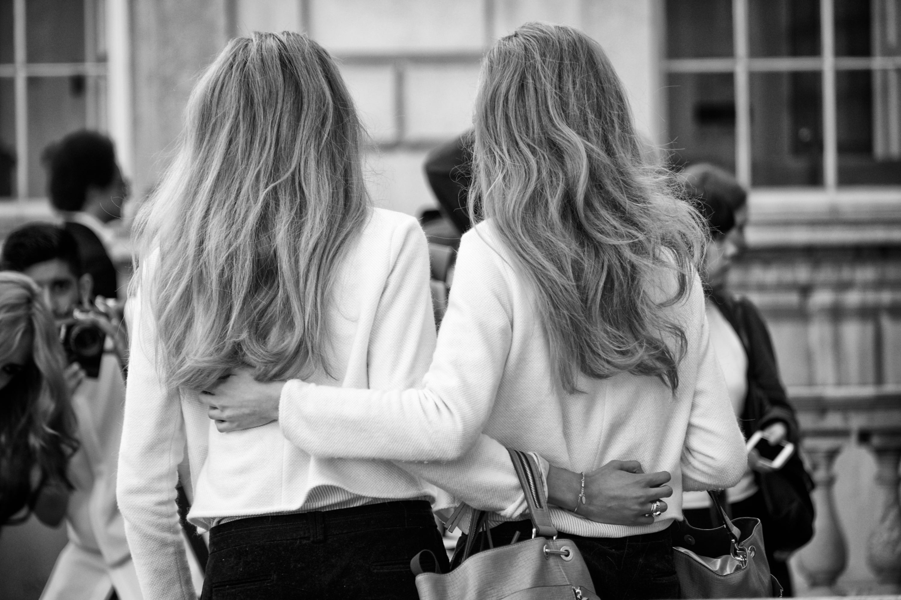 Black and white photograph of two women with long hair viewed from behind, wearing white sweaters, standing arm in arm outdoors.