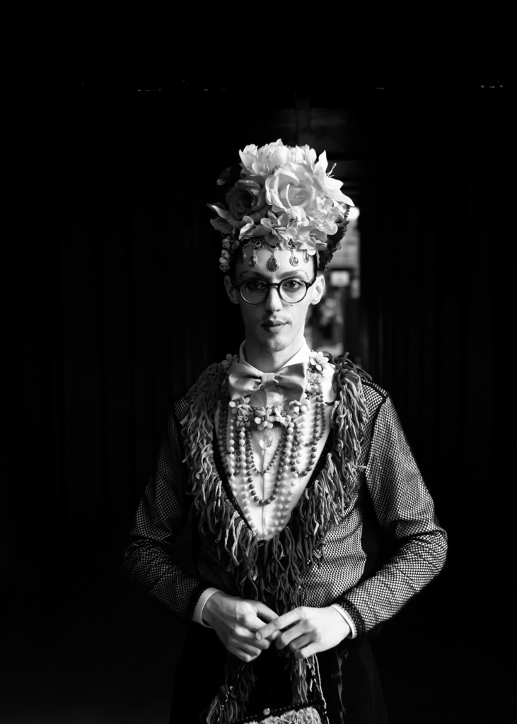 Black and white portrait of a person wearing oversized glasses, a large floral headdress, ornate jewelry, and patterned clothing against a dark background.