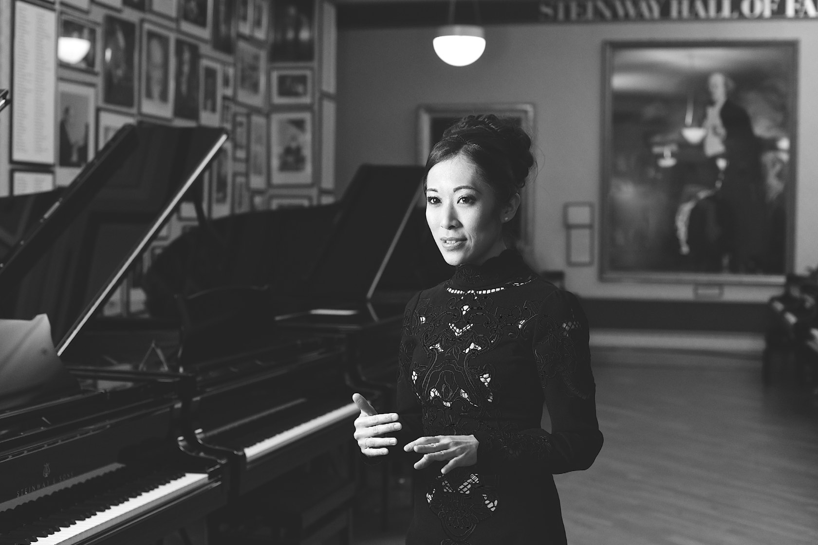 Black and white portrait of a woman in an ornate dark garment, standing near grand pianos in an elegant interior space.