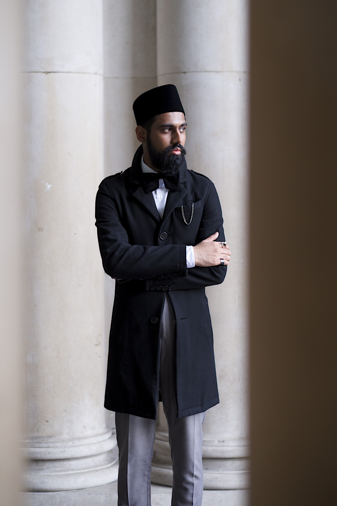 Man in formal black coat and traditional headwear stands between classical columns, arms crossed, gazing thoughtfully to the side.