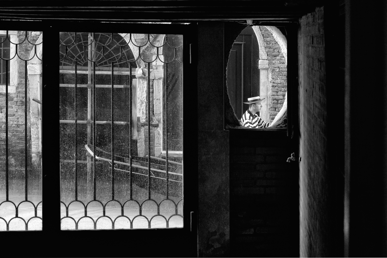 Black and white photograph showing a man in striped shirt and hat visible through a circular archway, with ornate iron gates in foreground
